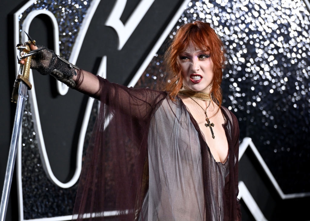 Person in sheer gown with a plunging neckline and gold choker holds a sword, posing at a formal event in front of a glittering backdrop