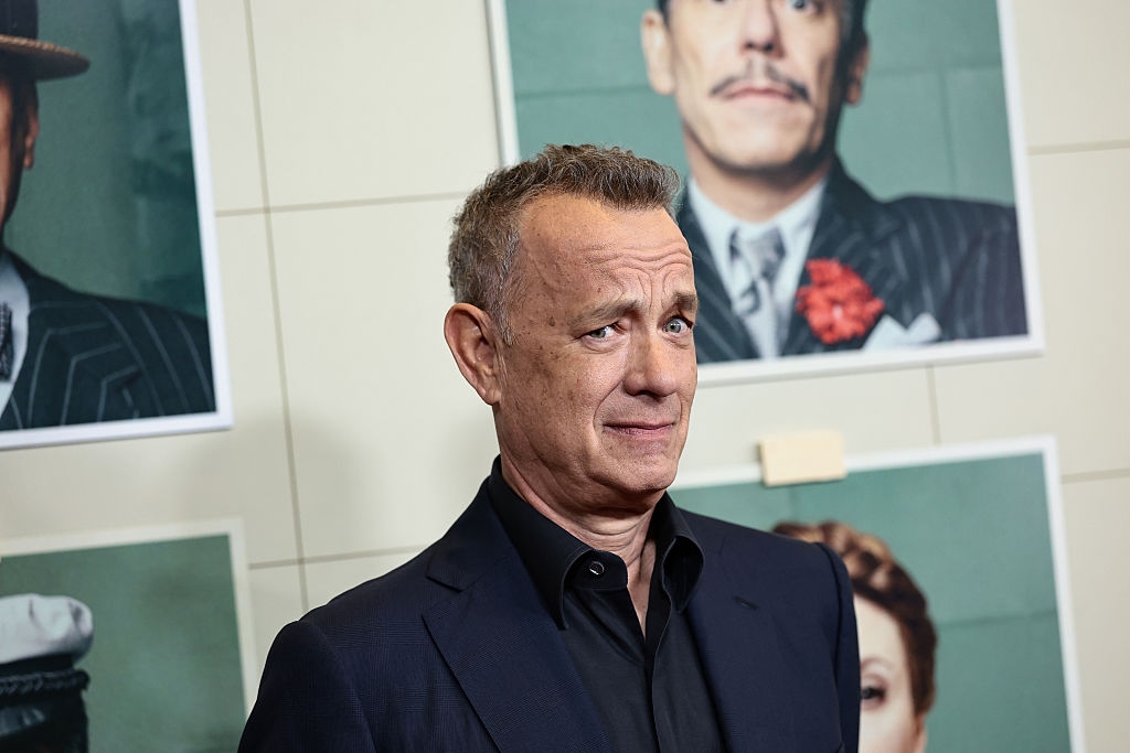 A person stands in front of vintage-style movie posters, wearing a dark suit and displaying a thoughtful expression