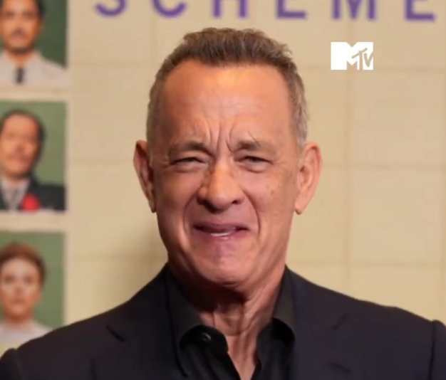 Person in a dark suit, smiling at an MTV event backdrop with small portraits