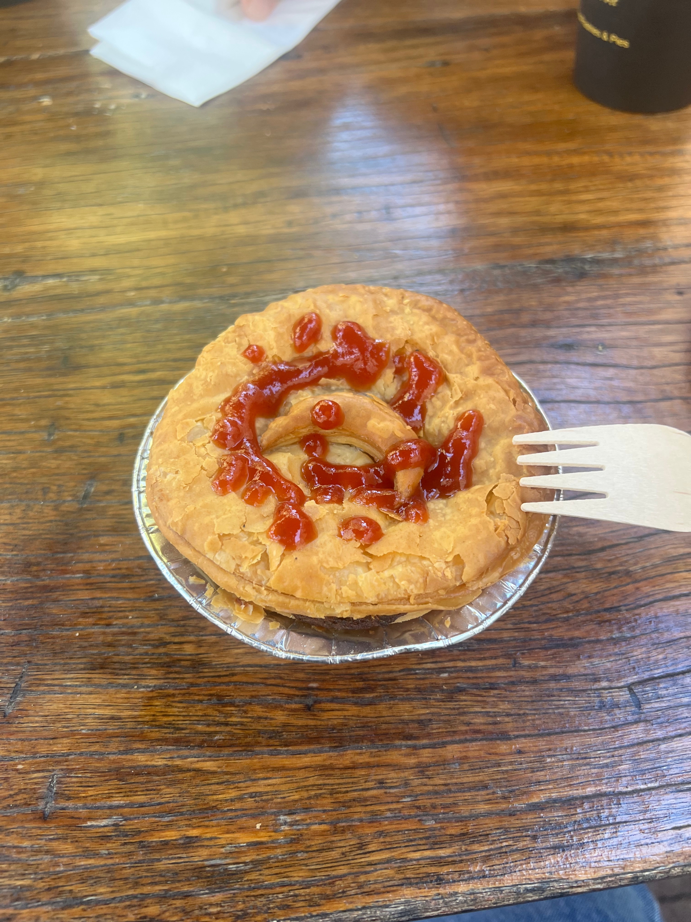 Meat pie with ketchup spiral on top, placed on a wooden table, with a wooden fork beside it