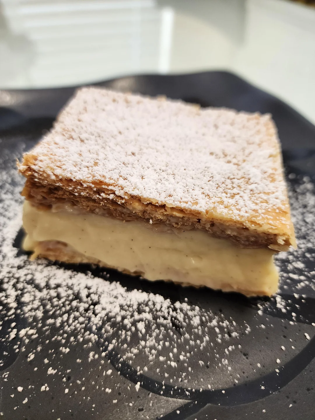 Pastry slice with flaky layers, creamy filling, and powdered sugar on top, served on a plate