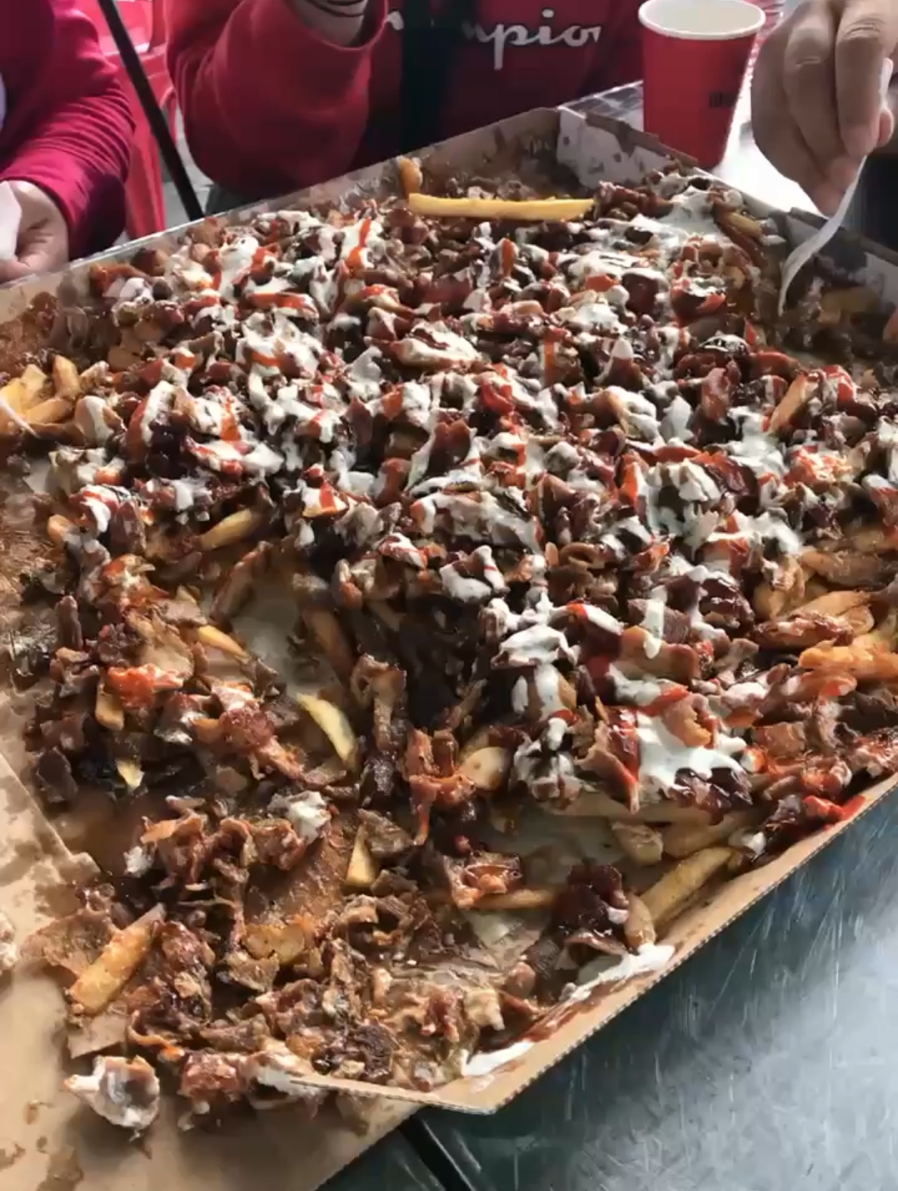 Large tray of loaded fries topped with meat, drizzled with sauce, being shared by a group with plastic forks