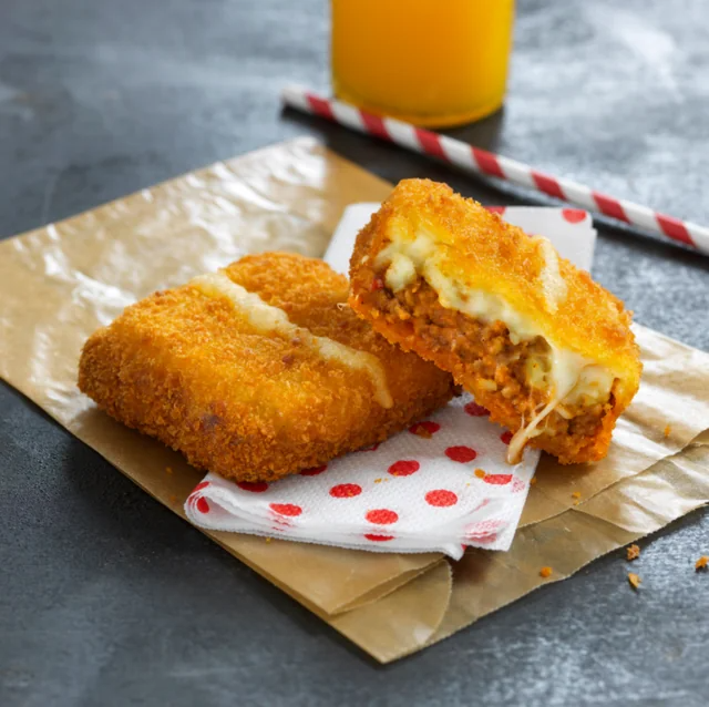 Breaded croquettes with cheese and ground meat filling on paper, next to a red polka dot napkin and a glass of juice with a striped straw