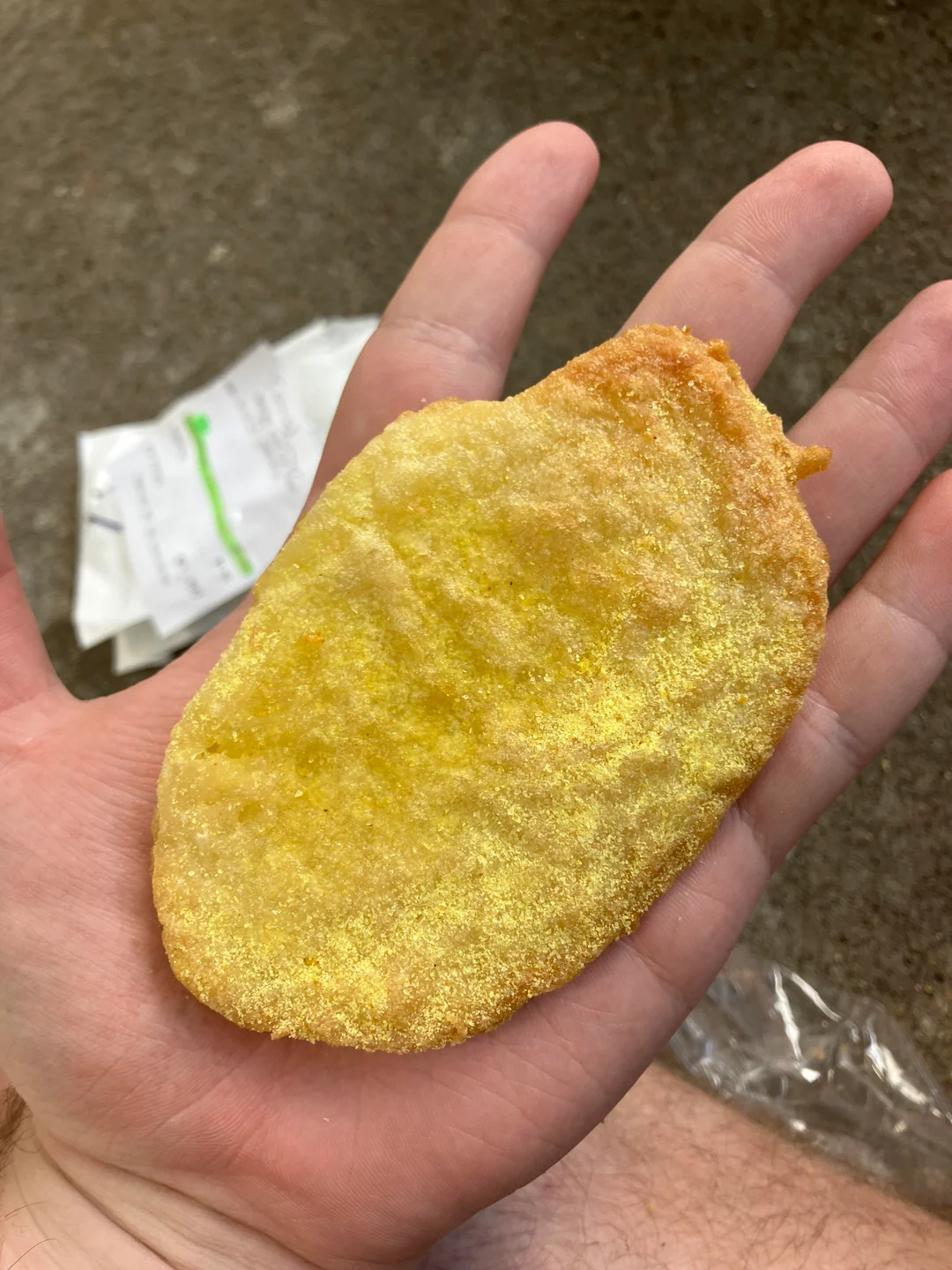 A large chip-like snack rests on a person's palm, showing its size relative to the hand. Packaging is visible in the background