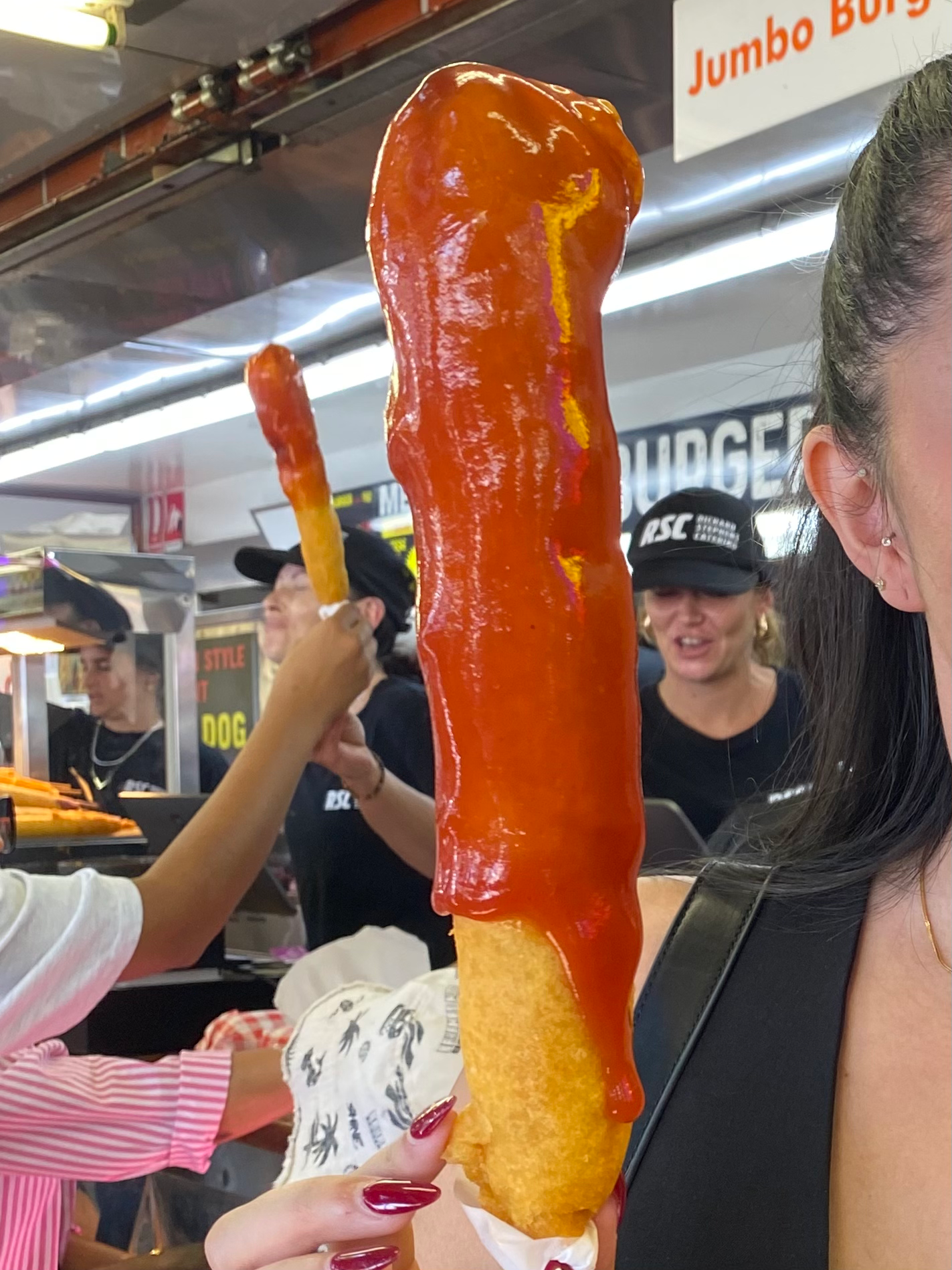 Close-up of a person holding a corn dog with bright red sauce at a busy food concession stand, staff working in the background