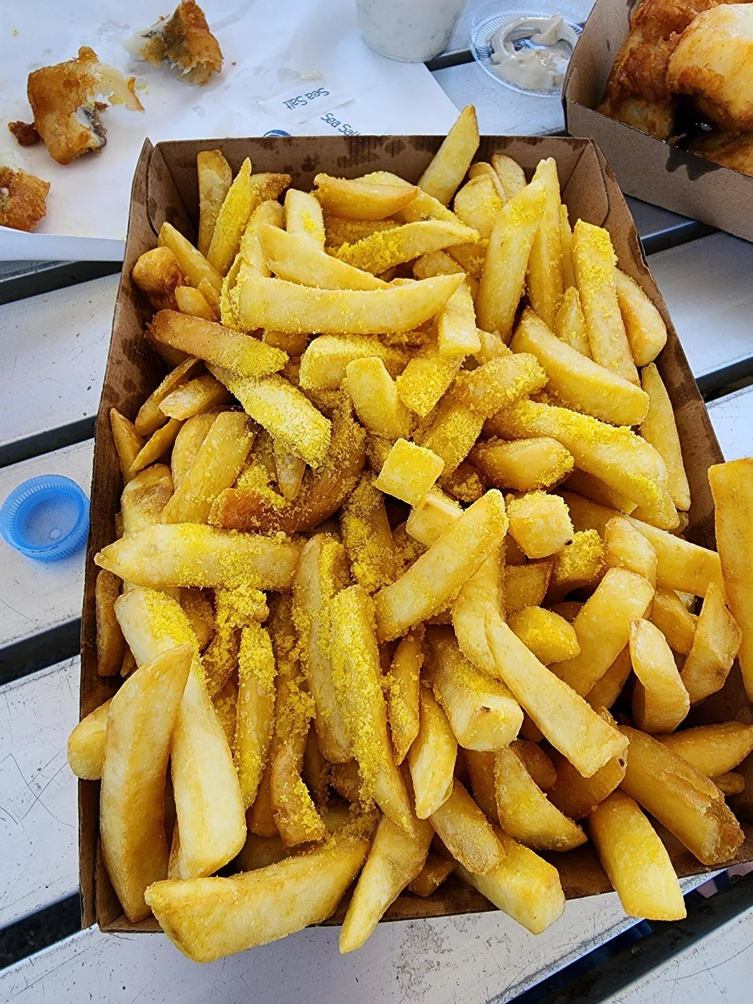 A tray filled with crispy fries, sprinkled with a generous amount of seasoning, sits on a table, with some food partially visible in the background