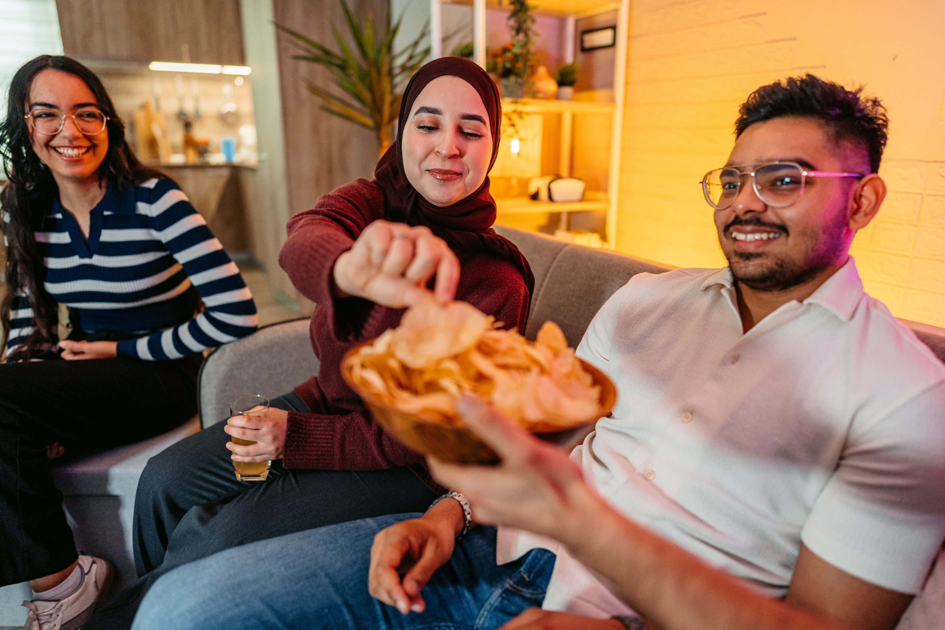 Tre venner sitter på en sofa og koser seg med snacks. En kvinne strekker seg etter chips, mens de alle smiler og engasjerer seg i tilfeldig samtale
