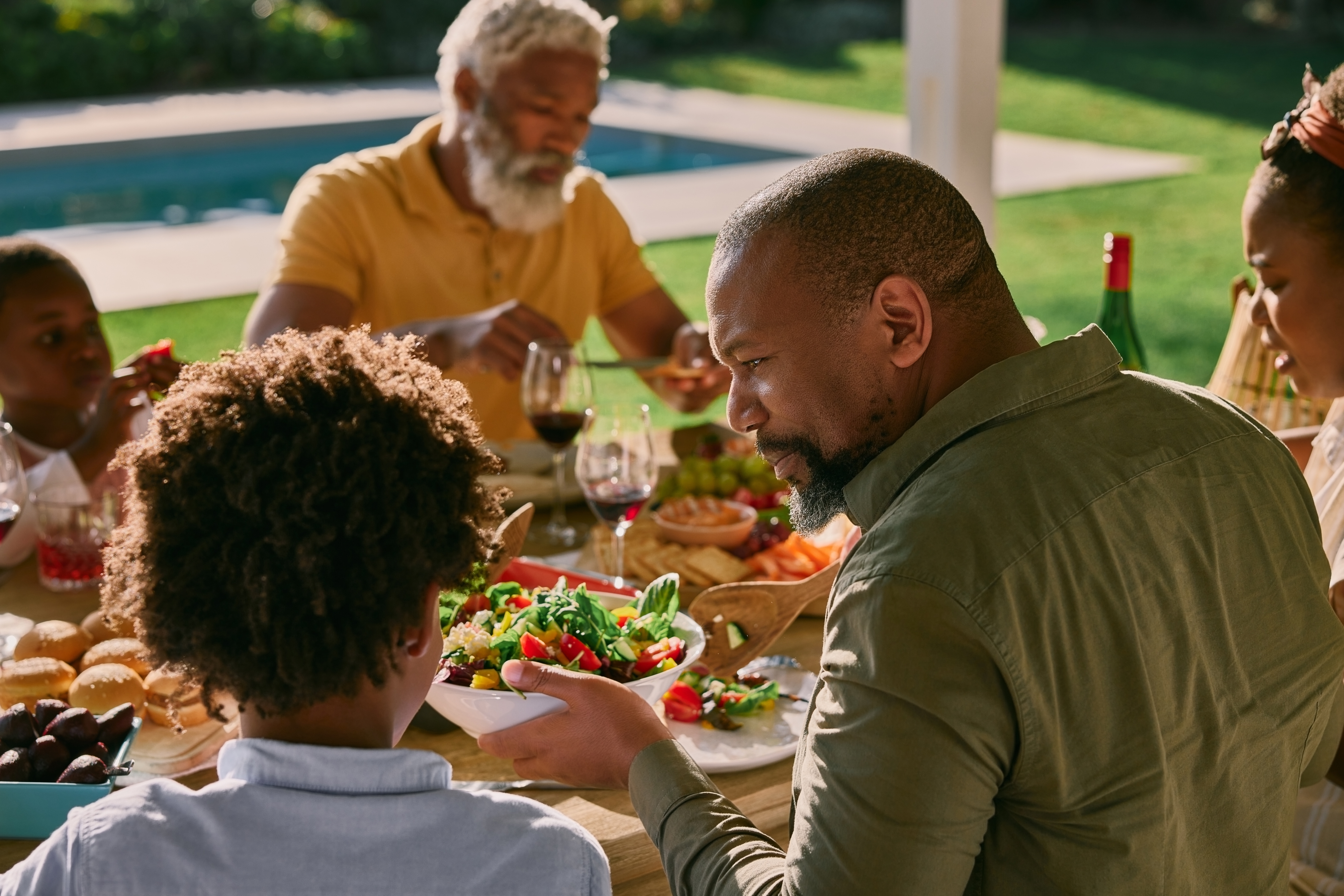 Familie som spiser og snakker ved et utendørsbord, med en mann som serverer salat til et barn. En hage og basseng er i bakgrunnen