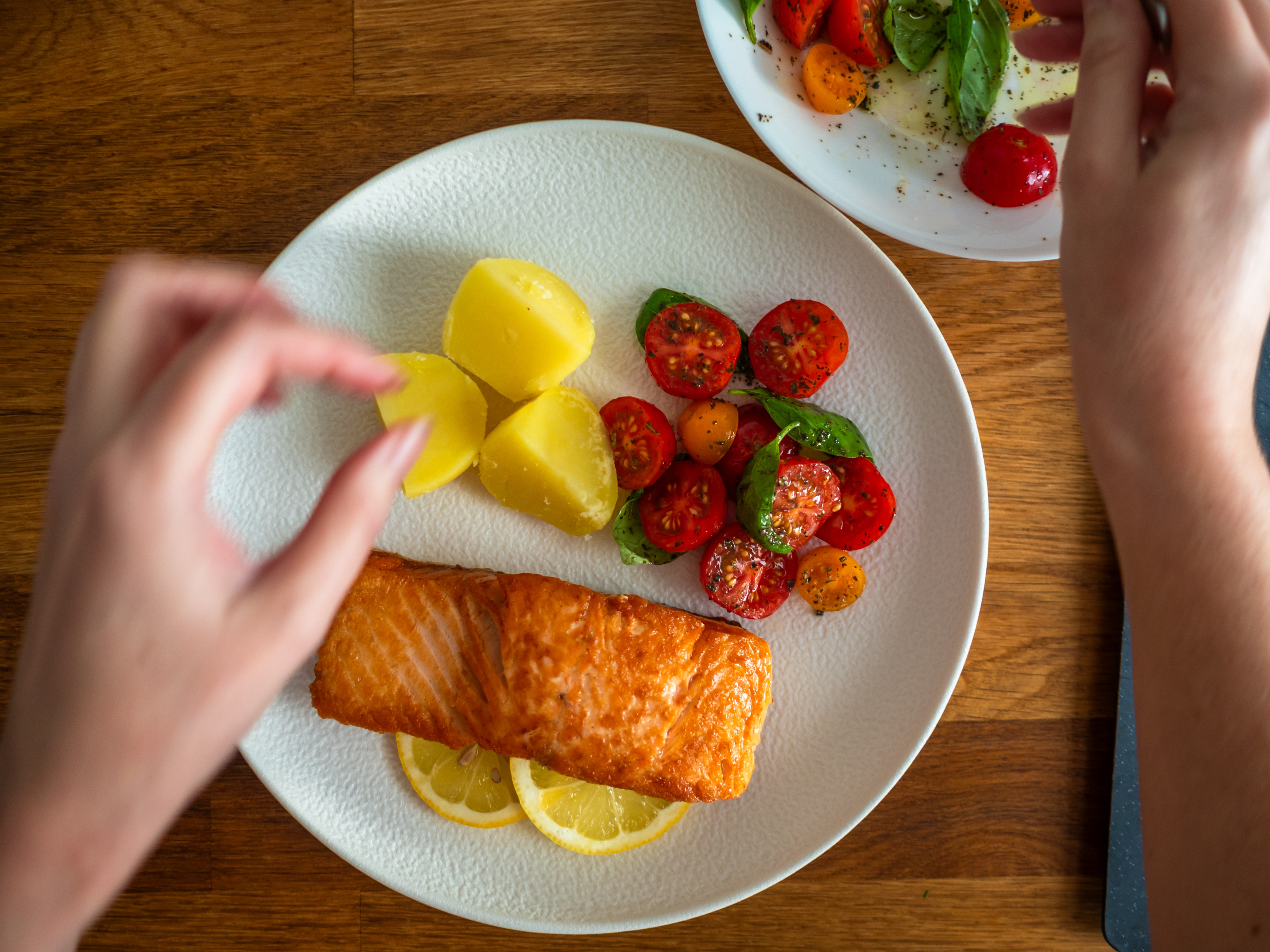 Laksefilet med sitronskiver, kirsebærtomater og poteter på en hvit tallerken; hender justerer matopplegget