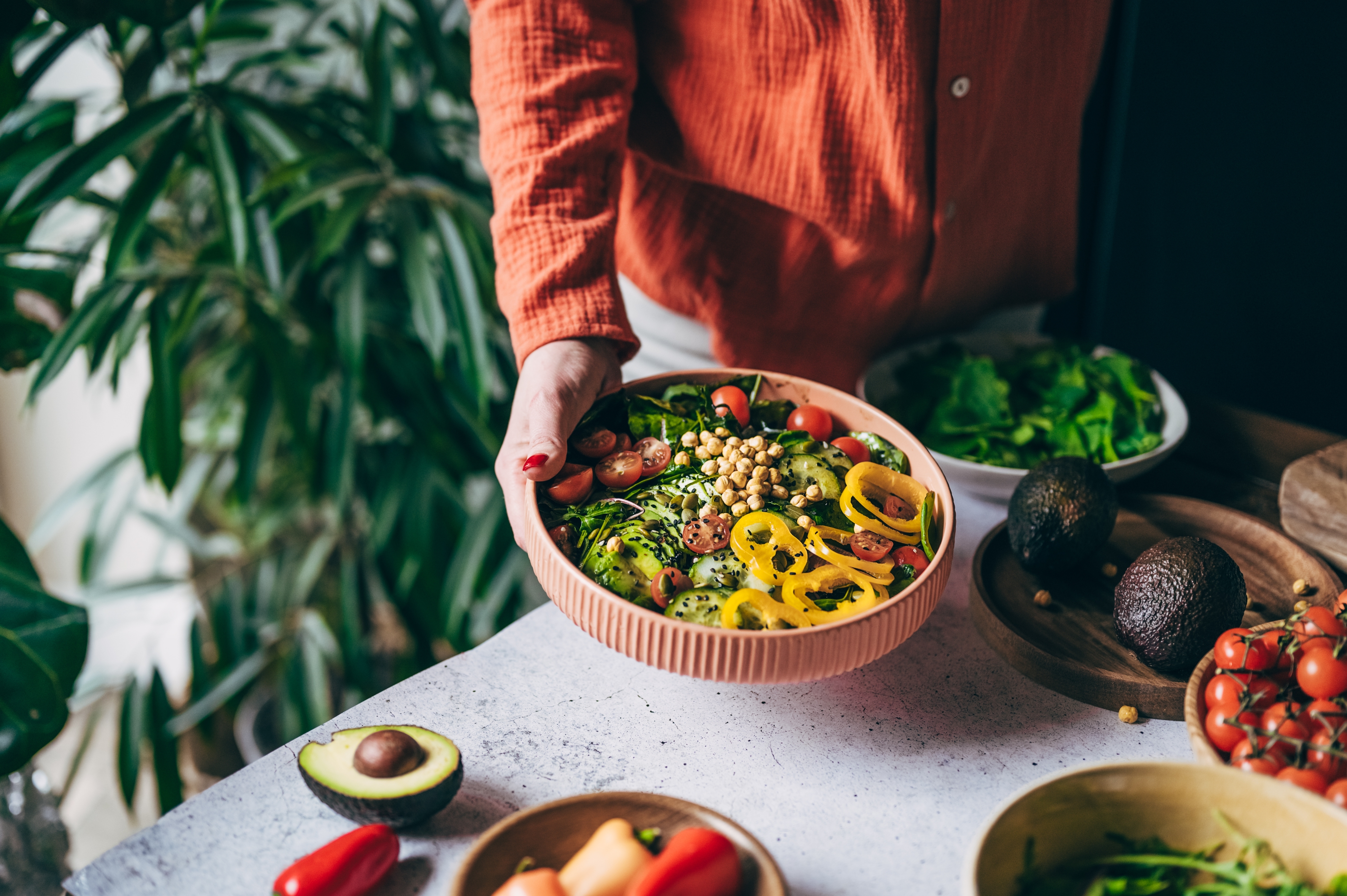 Person som holder en fargerik salatskål med tomater, mais og paprika, omgitt av ferske ingredienser og planter