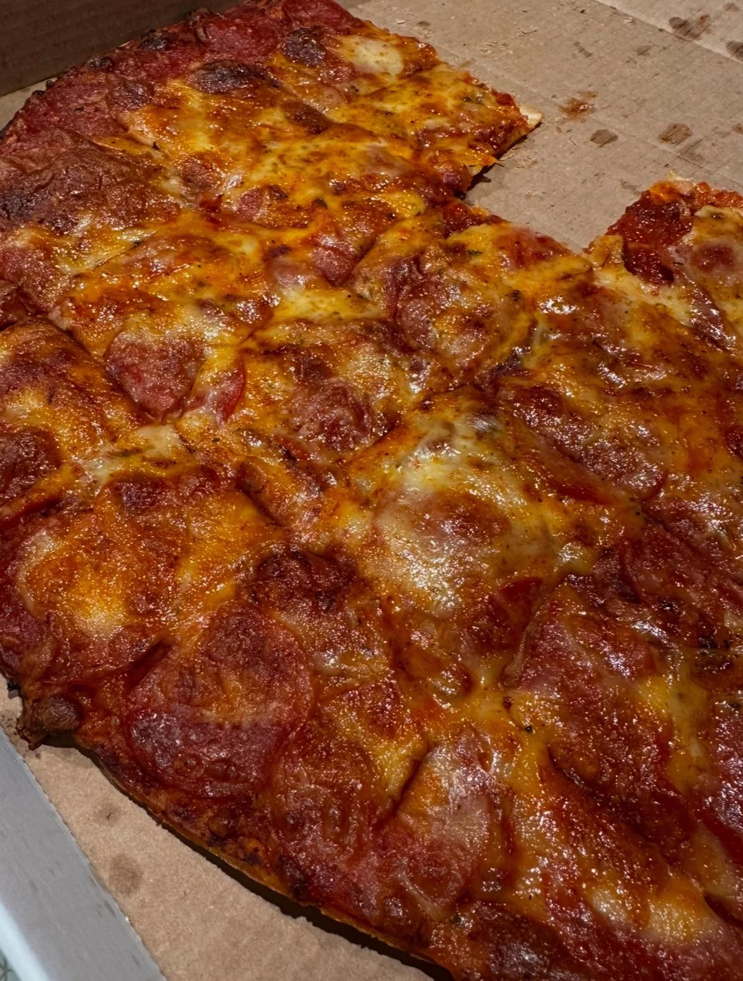 Pizza with pepperoni and cheese, cut into squares, sits on a cardboard surface. Some slices are missing from the large portion