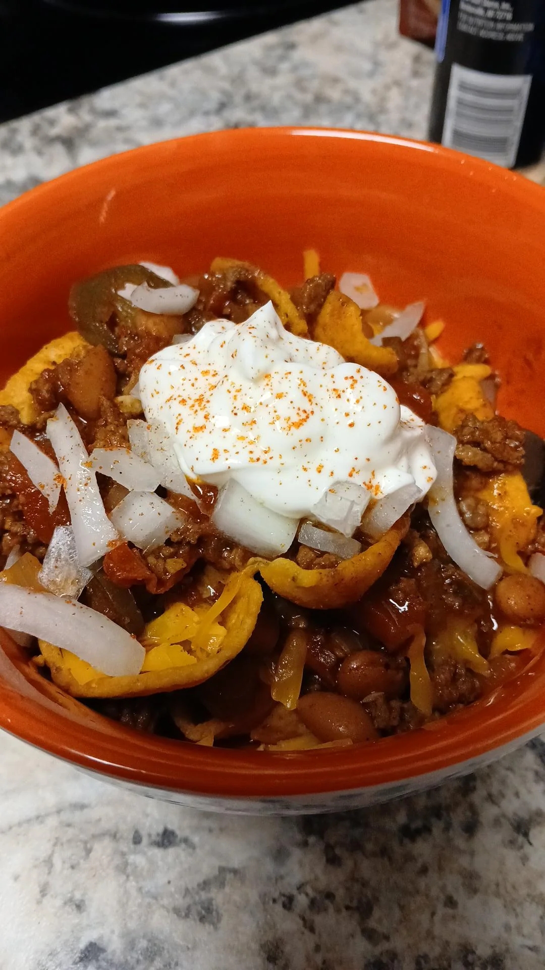 A bowl of chili with ground meat, beans, cheese, onions, and a dollop of sour cream, sprinkled with seasoning