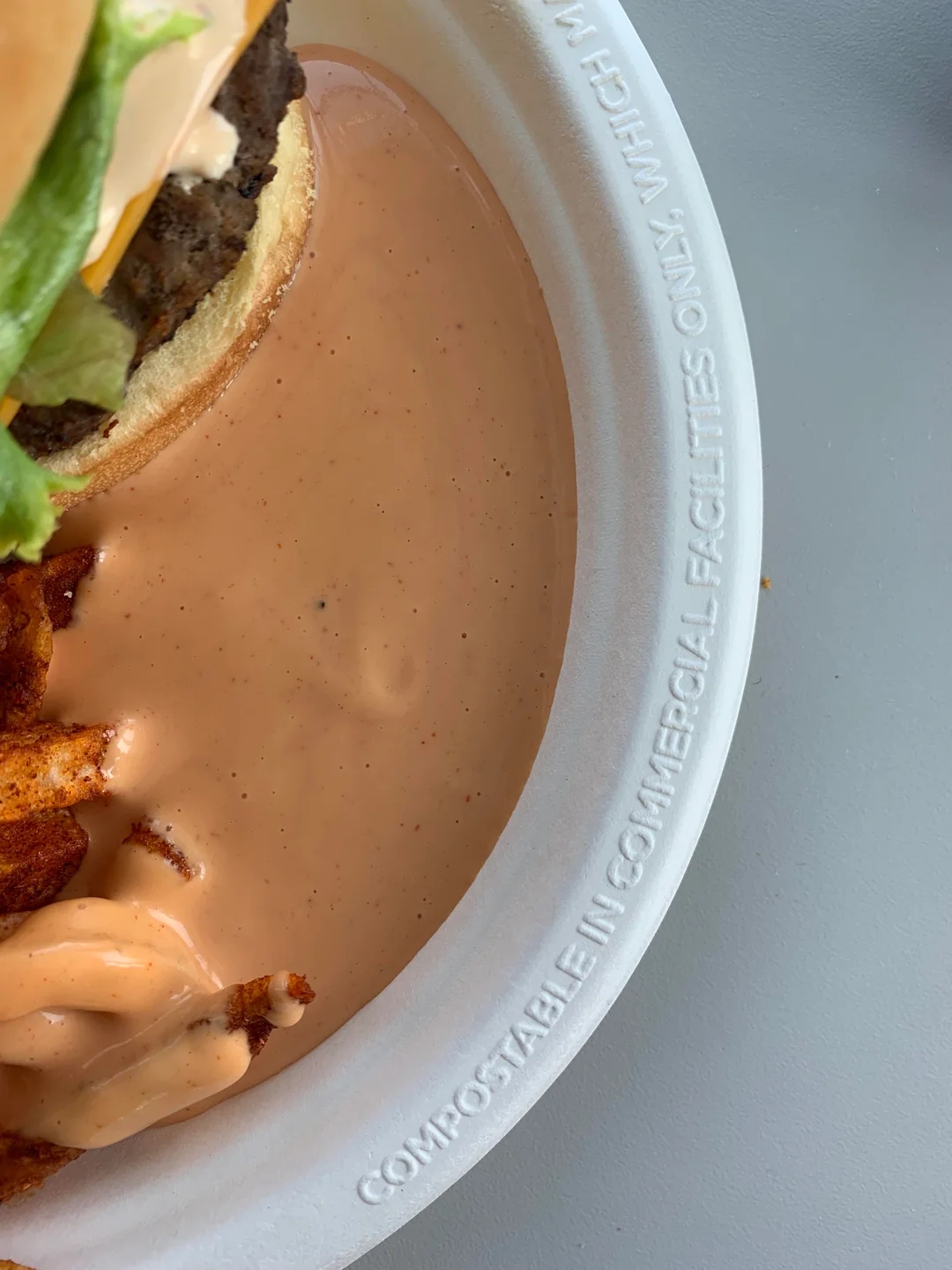 Burger with lettuce and cheese on a bun, next to waffle fries and a creamy sauce on a disposable plate. Plate is compostable