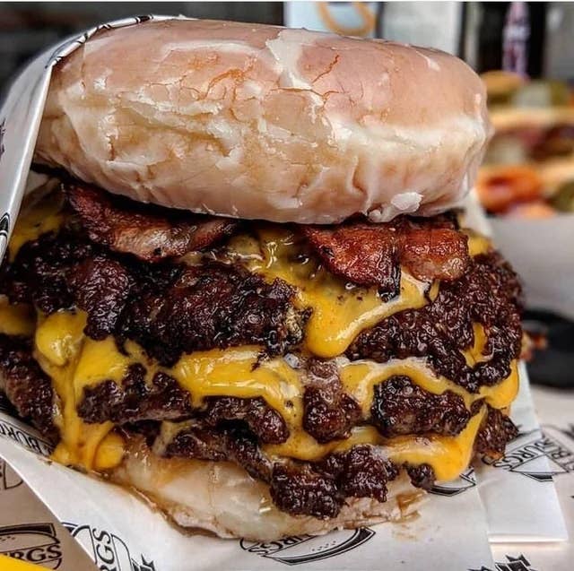Massive burger with multiple beef patties, melted cheese, crispy bacon, and a glazed donut as a bun, wrapped in branded paper