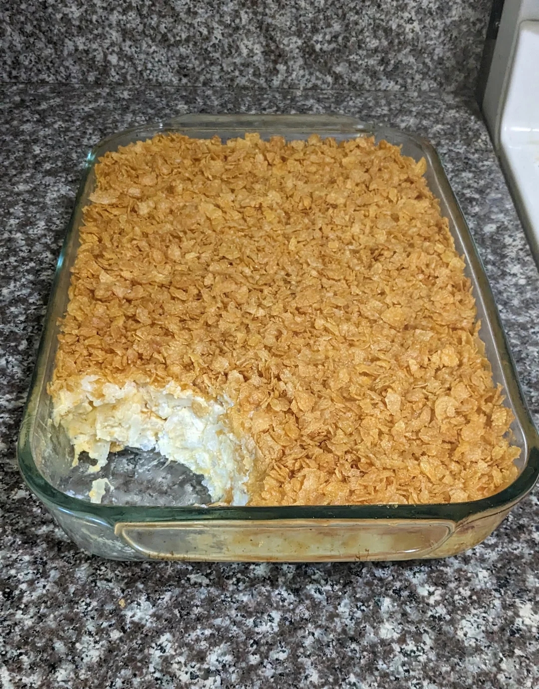 Casserole dish with creamy interior and crunchy flake topping, with a portion missing. Placed on a granite countertop
