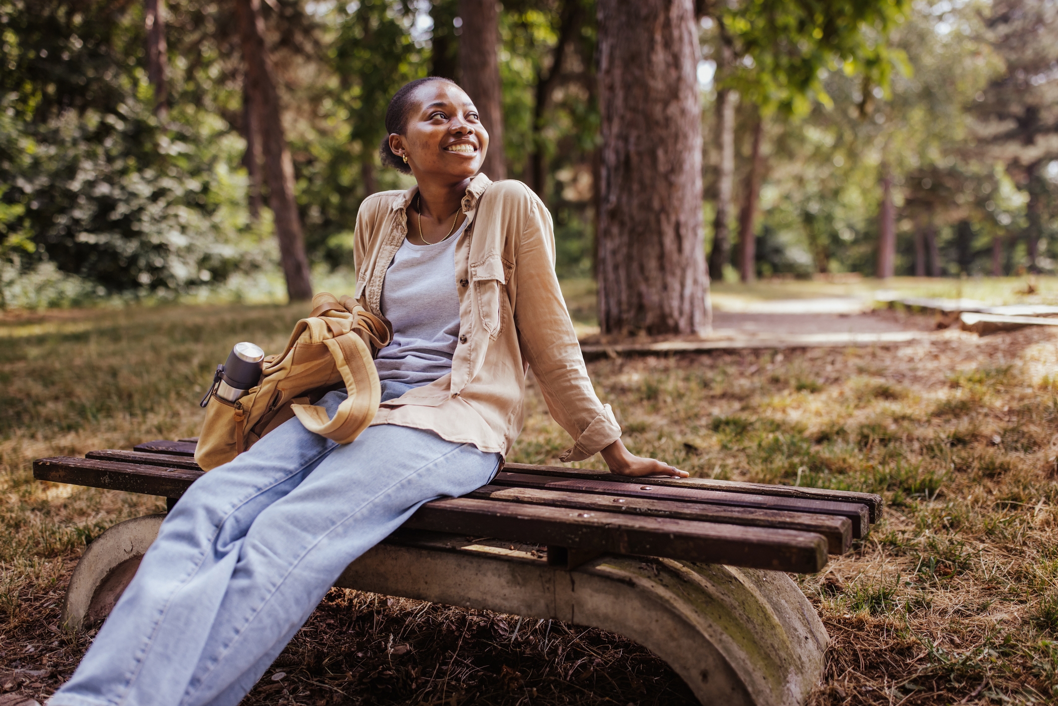 Person slappet av på en parkbenk, smilende, iført tilfeldige jeans og en lysjakke, omgitt av trær