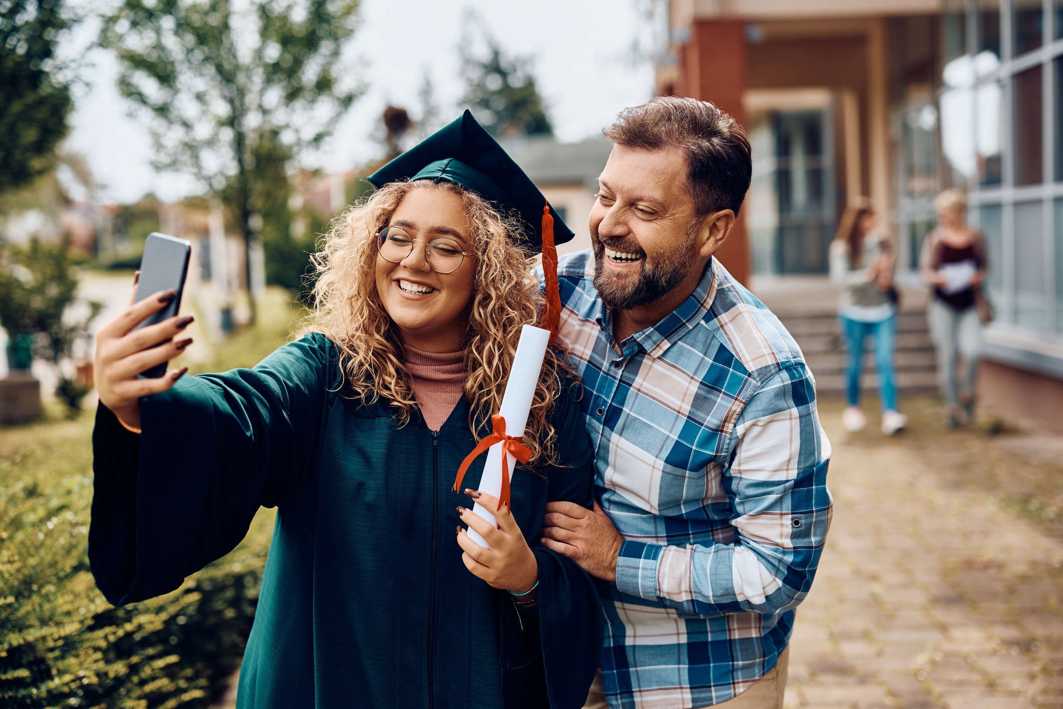 Graduate in Cap and Gown Taking a Selfie med en eldre mann, begge smilende gledelig utendørs, noe som antyder en feiring av en akademisk prestasjon
