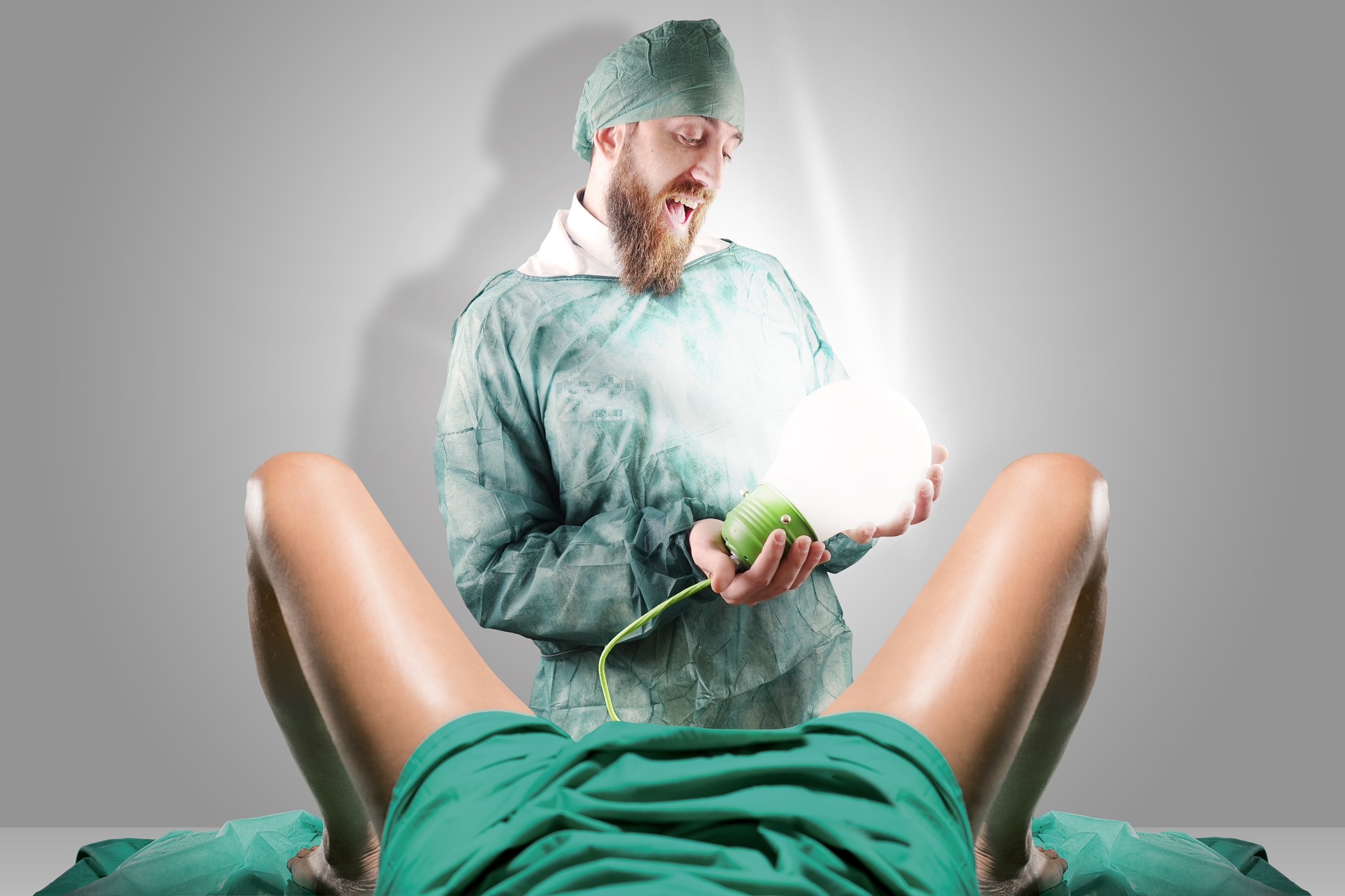 A bearded person in surgical attire humorously holds a glowing bulb instead of a newborn during a comical delivery scene