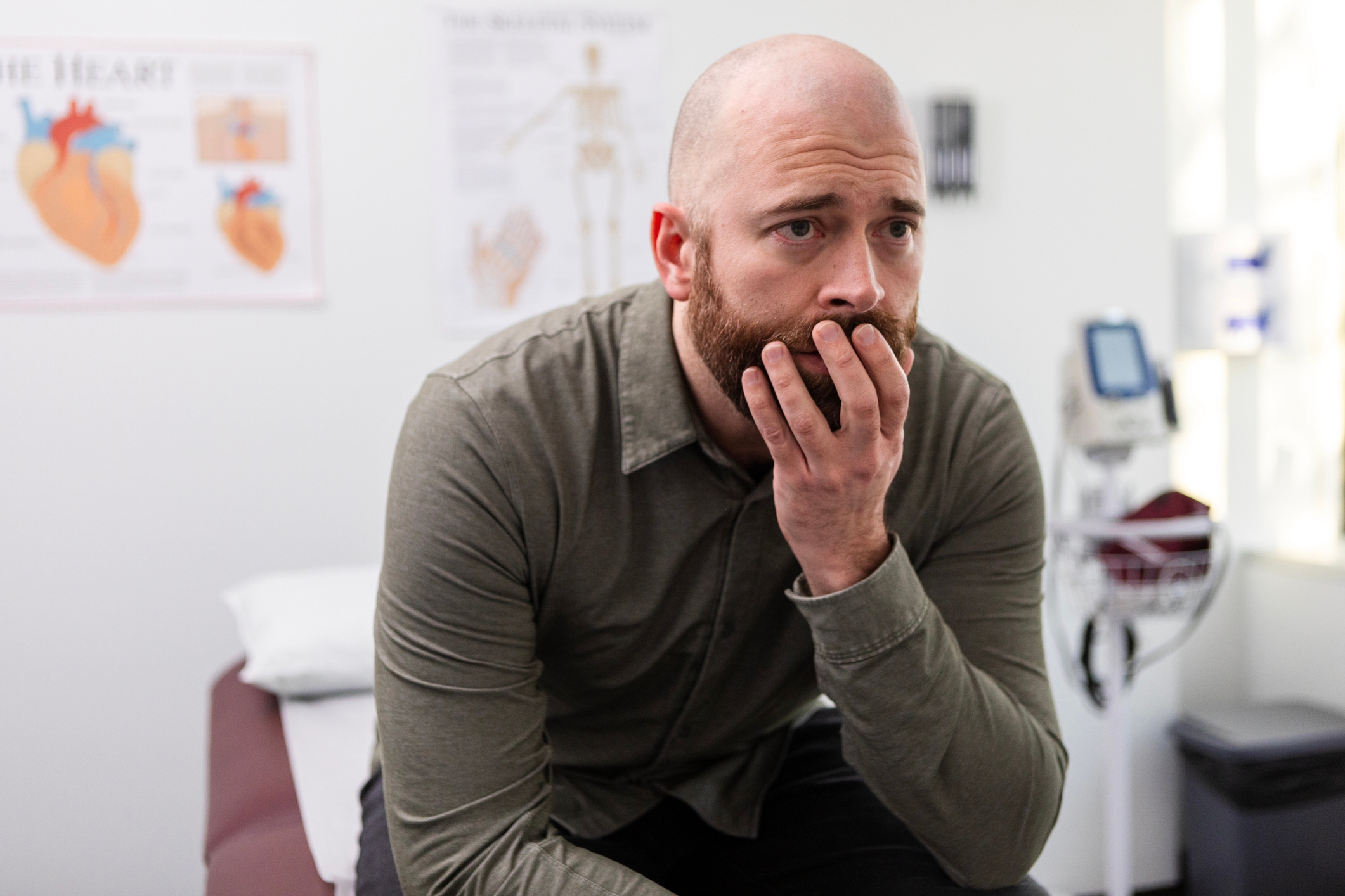 A worried man sits in a doctor&#x27;s office, hand to mouth, with medical posters and equipment in the background