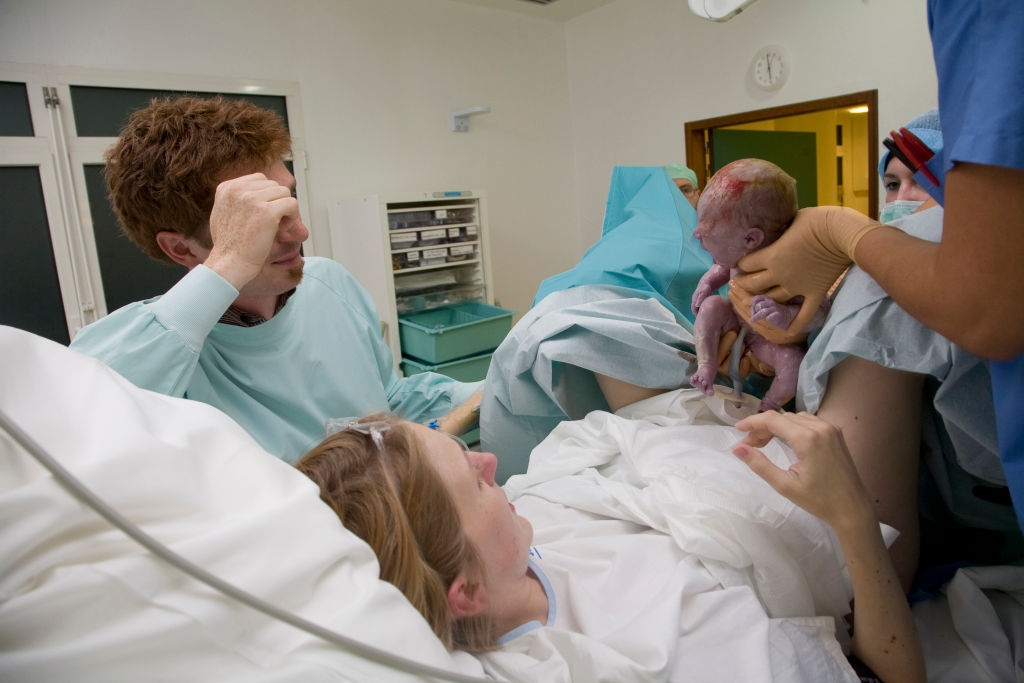 A new mother lies in a hospital bed, looking at a nurse holding her newborn. A man sits beside her, smiling at the baby
