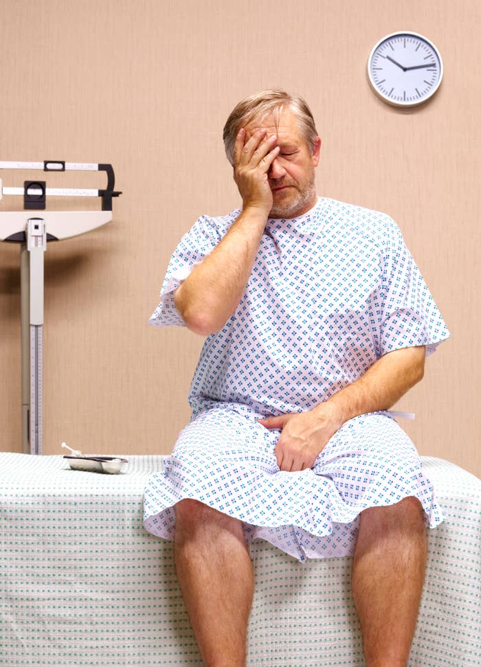 A man in a hospital gown looks distressed, sitting on an examination table beside medical equipment, with a clock on the wall