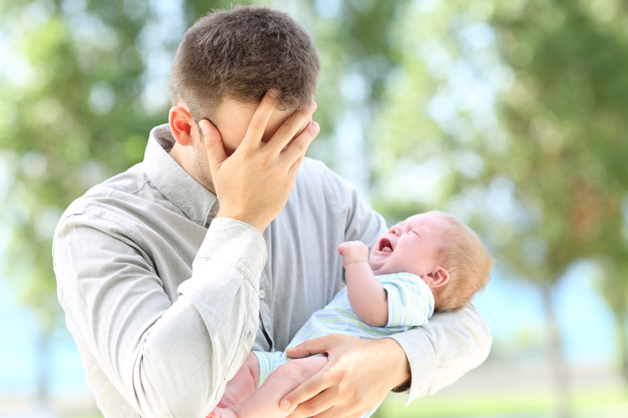 A man holds a crying baby, covering his face with his hand, possibly feeling stressed or overwhelmed
