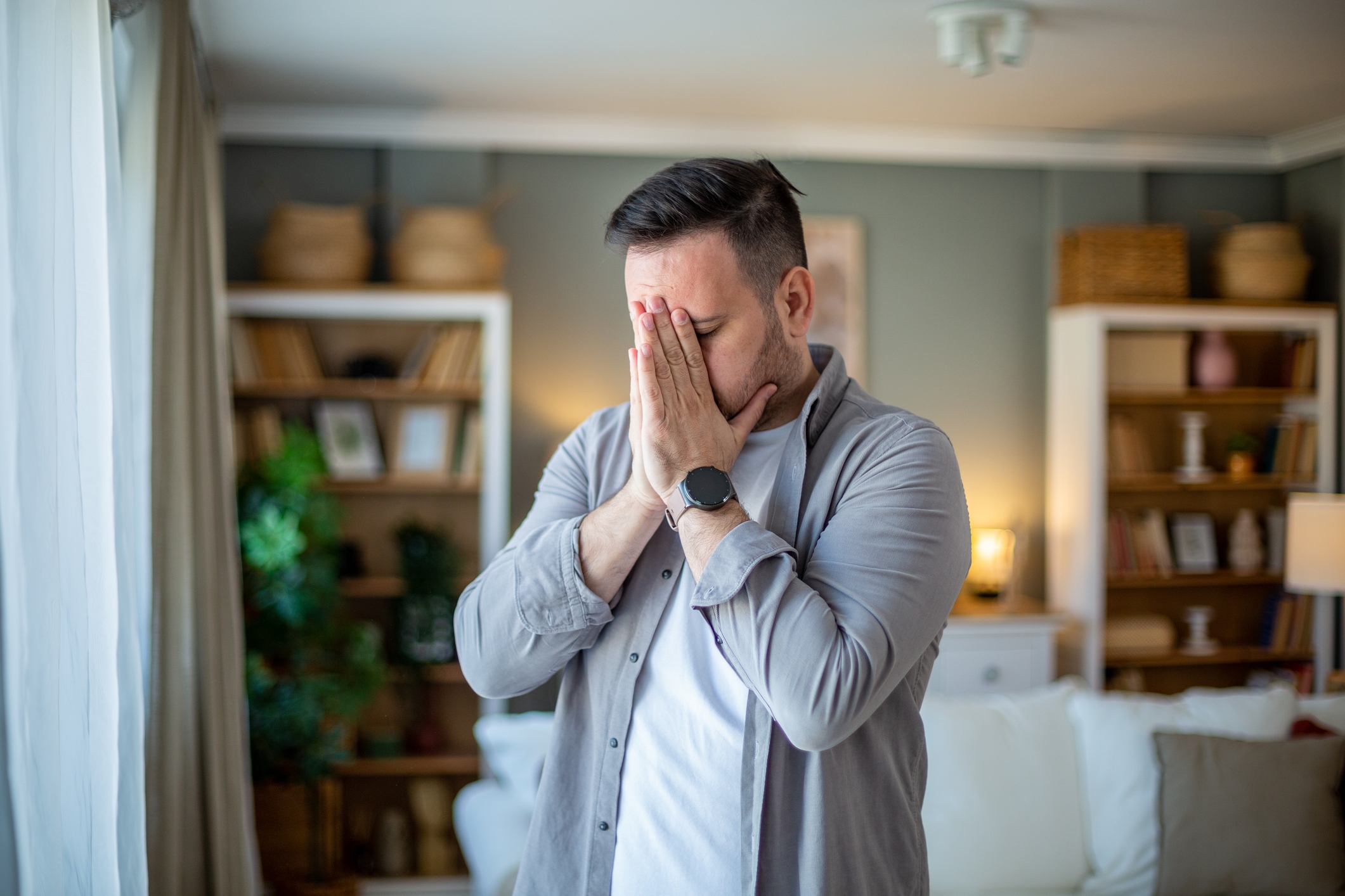 Person in a casual outfit stands in a living room, appearing thoughtful with hands clasped near face, surrounded by bookshelves and plants