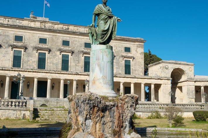 ギリシャの宮殿前に立つクラシックな彫像のディティール。背景に歴史的建物と青空。