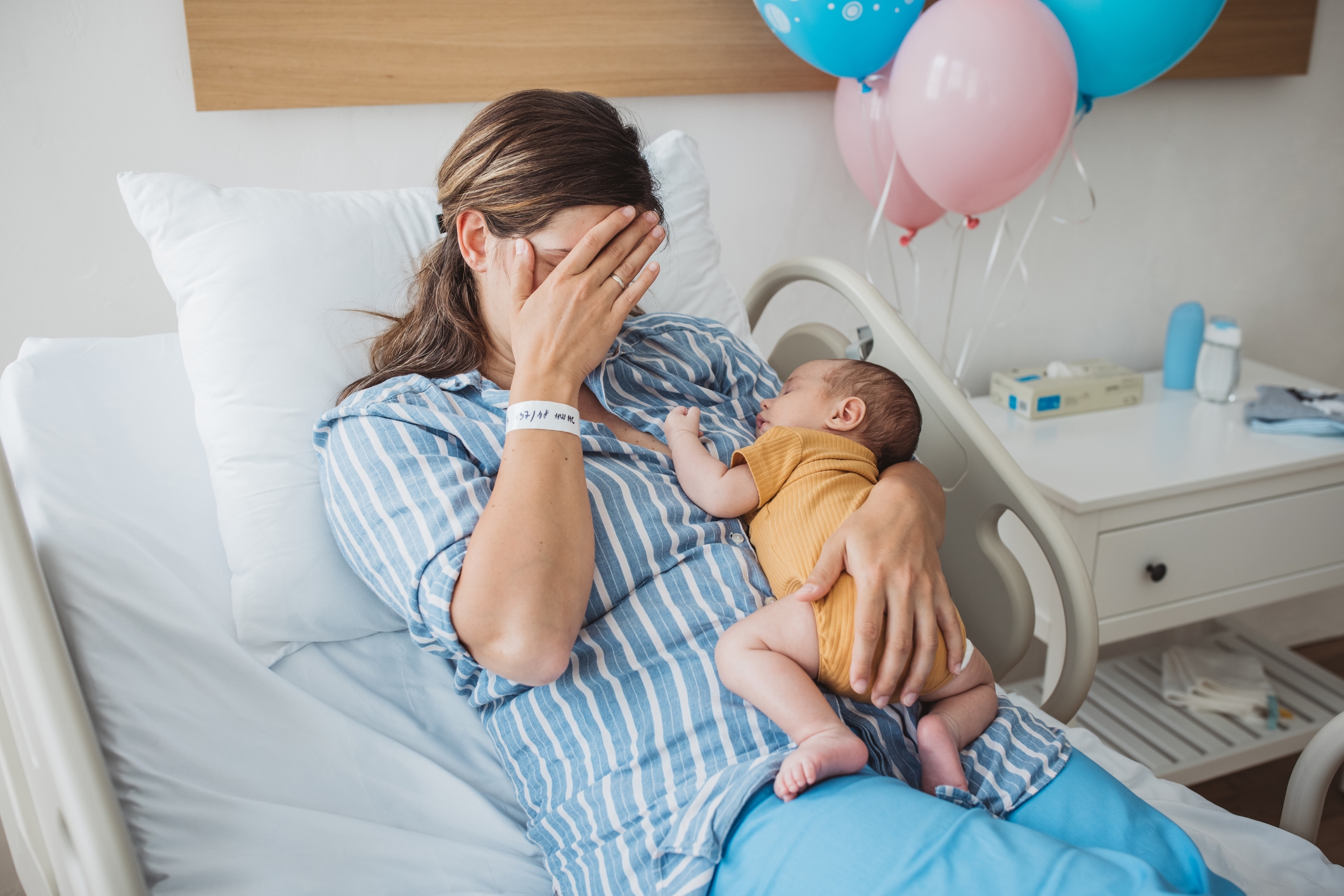 Person in a hospital bed, wearing striped pajamas, holds a baby and covers their face with one hand. Balloons are in the background