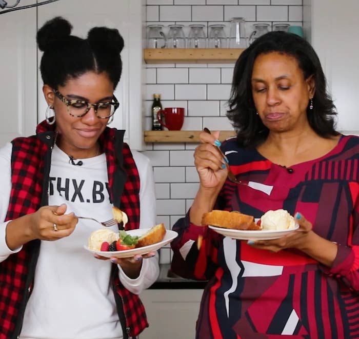 Two people enjoying cake with ice cream in a kitchen, wearing casual attire. Shelves with kitchen items are visible in the background