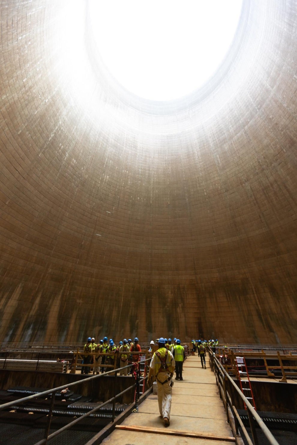 Workers in safety gear walk along a platform inside a large, industrial cooling tower, looking up towards the open top
