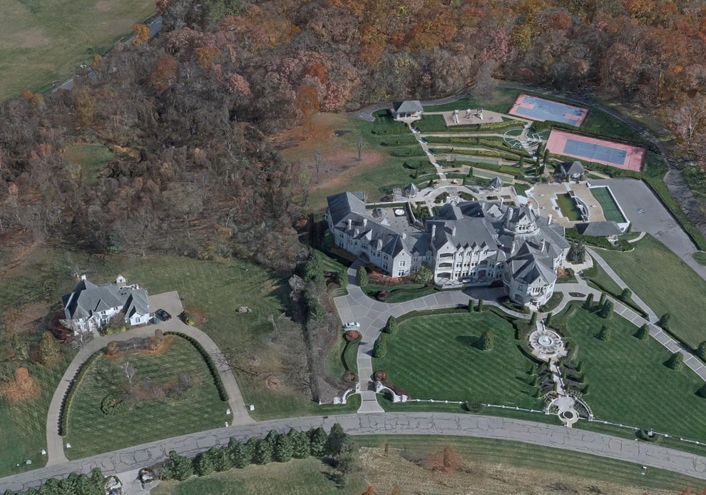 Aerial view of a large, ornate mansion with manicured gardens, surrounded by trees and several tennis courts nearby