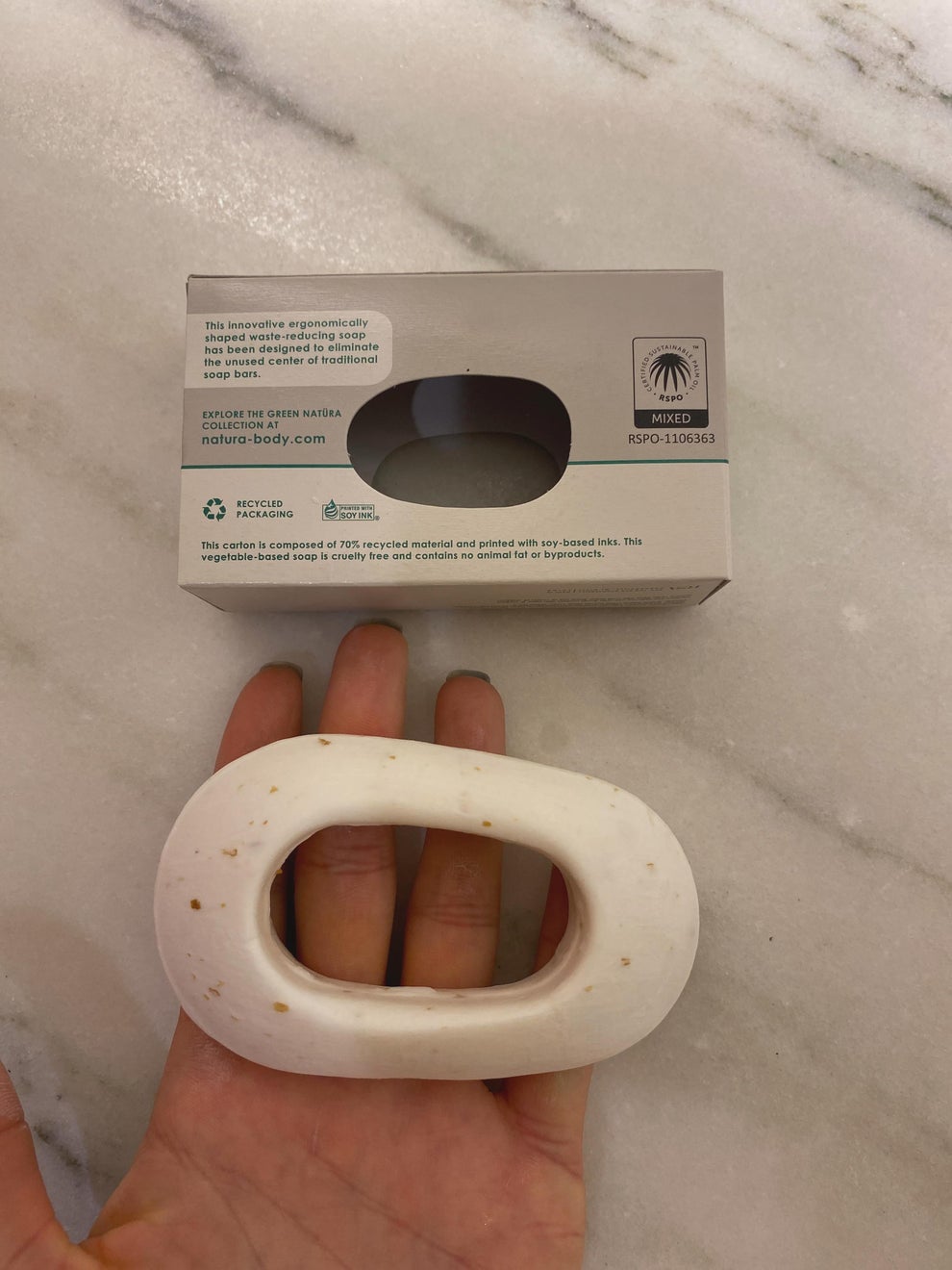 Unique ergonomic soap shaped as an oval ring placed on a hand, with eco-friendly packaging in the background