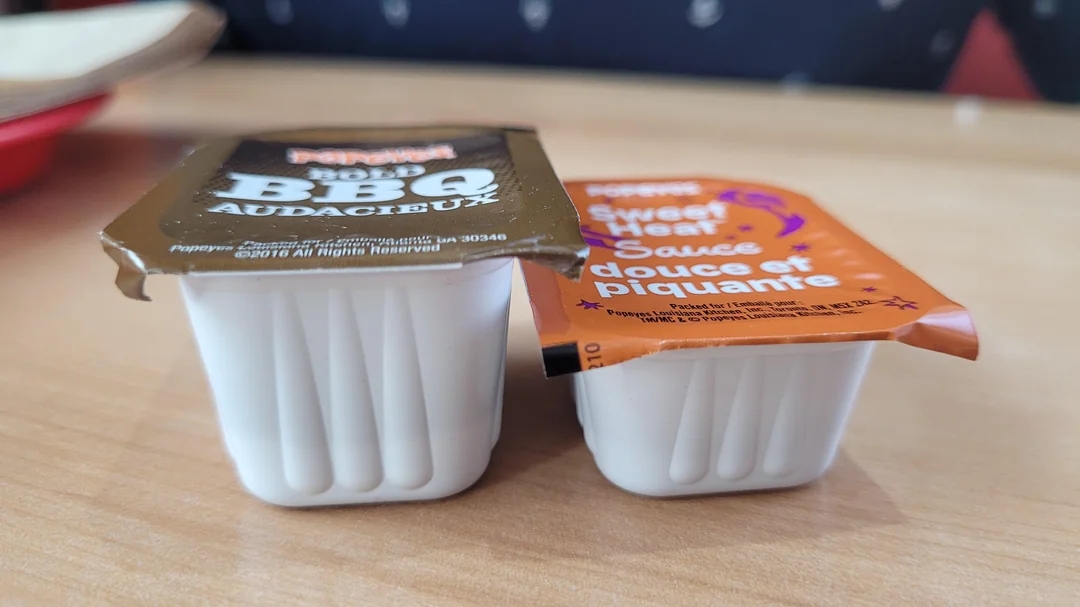 Two fast food sauce packets sit on a table: BBQ and Sweet &amp;amp; Sour. Both packets are white with peelable labels on top