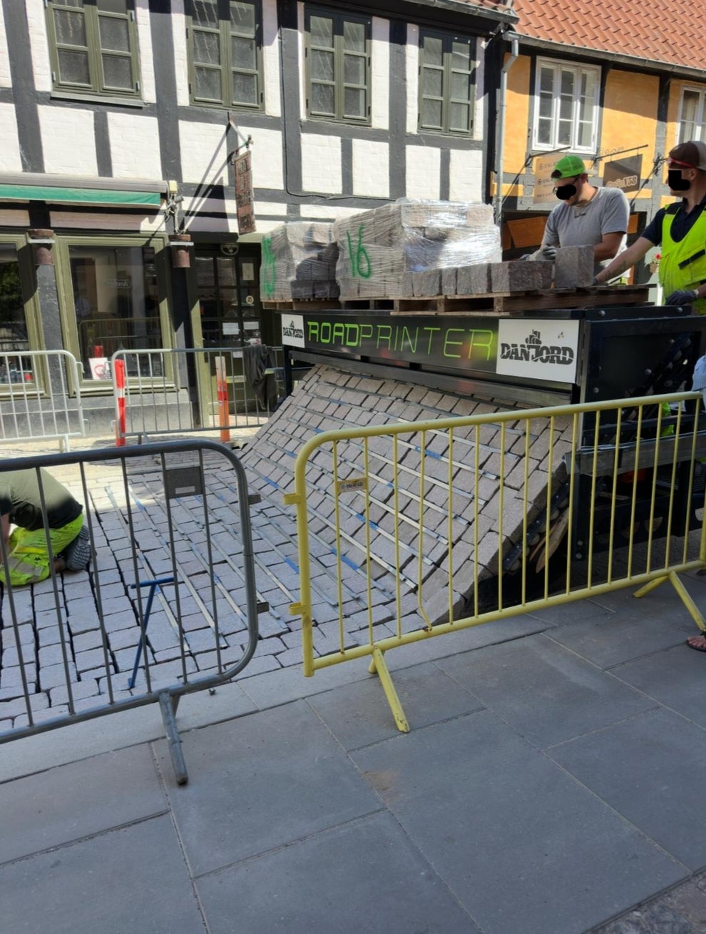 Machine laying brick road with workers supervising in a city setting; yellow barriers surround the area