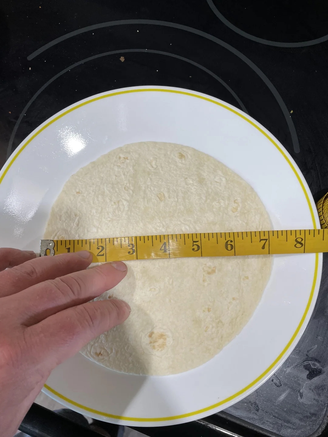 A hand holding a measuring tape across a tortilla on a plate, measuring its diameter at 7 inches