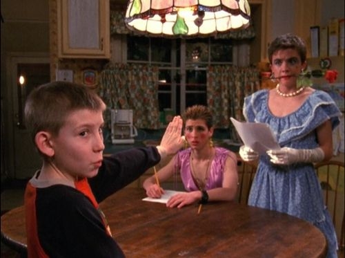 Three people in quirky attire at a kitchen table; a boy raises his hand, an adult in a ruffled dress holds papers, and another wears a pink top