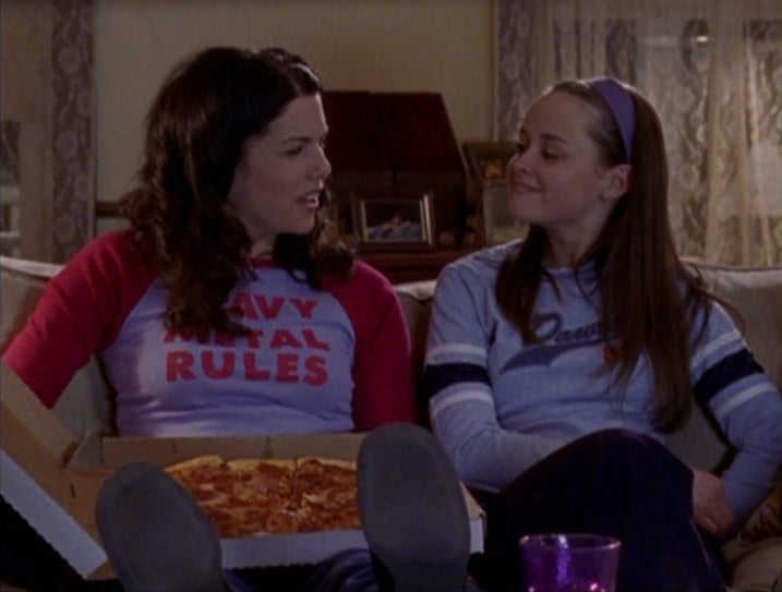 Two people sitting on a couch with a pizza box. One wears a "Heavy Metal Rules" shirt; both seem to be engaged in conversation, smiling slightly