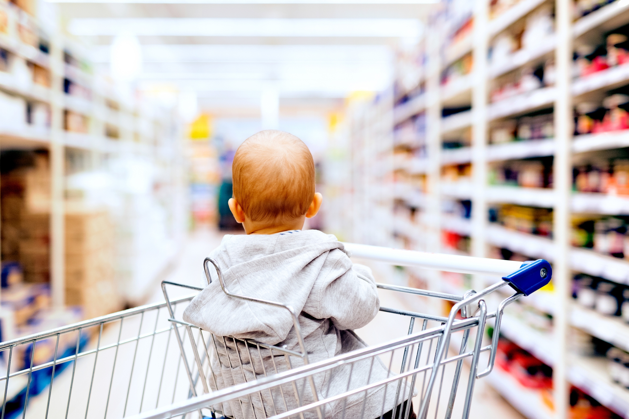 Baby i en handlekurv som ser på hyller i matbutikker, iført hettejakke