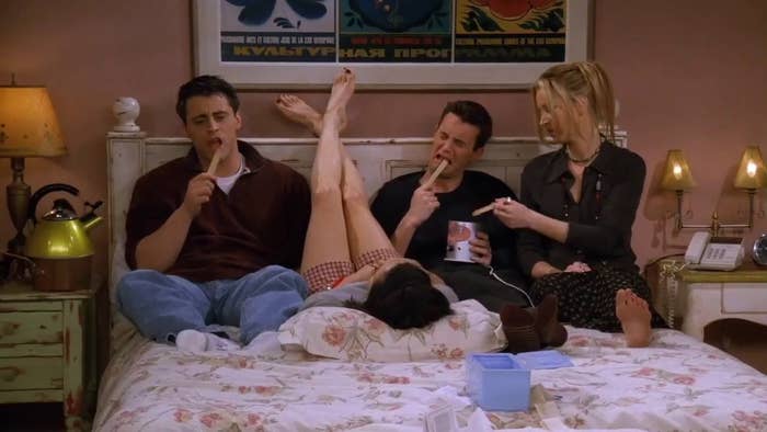 Three friends sit on a bed, enjoying ice cream, while a fourth person lies with legs raised. It's a casual and relaxed scene from a TV show