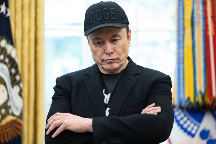 Elon Musk wearing a "Doge" cap and black blazer stands with arms crossed, appearing contemplative