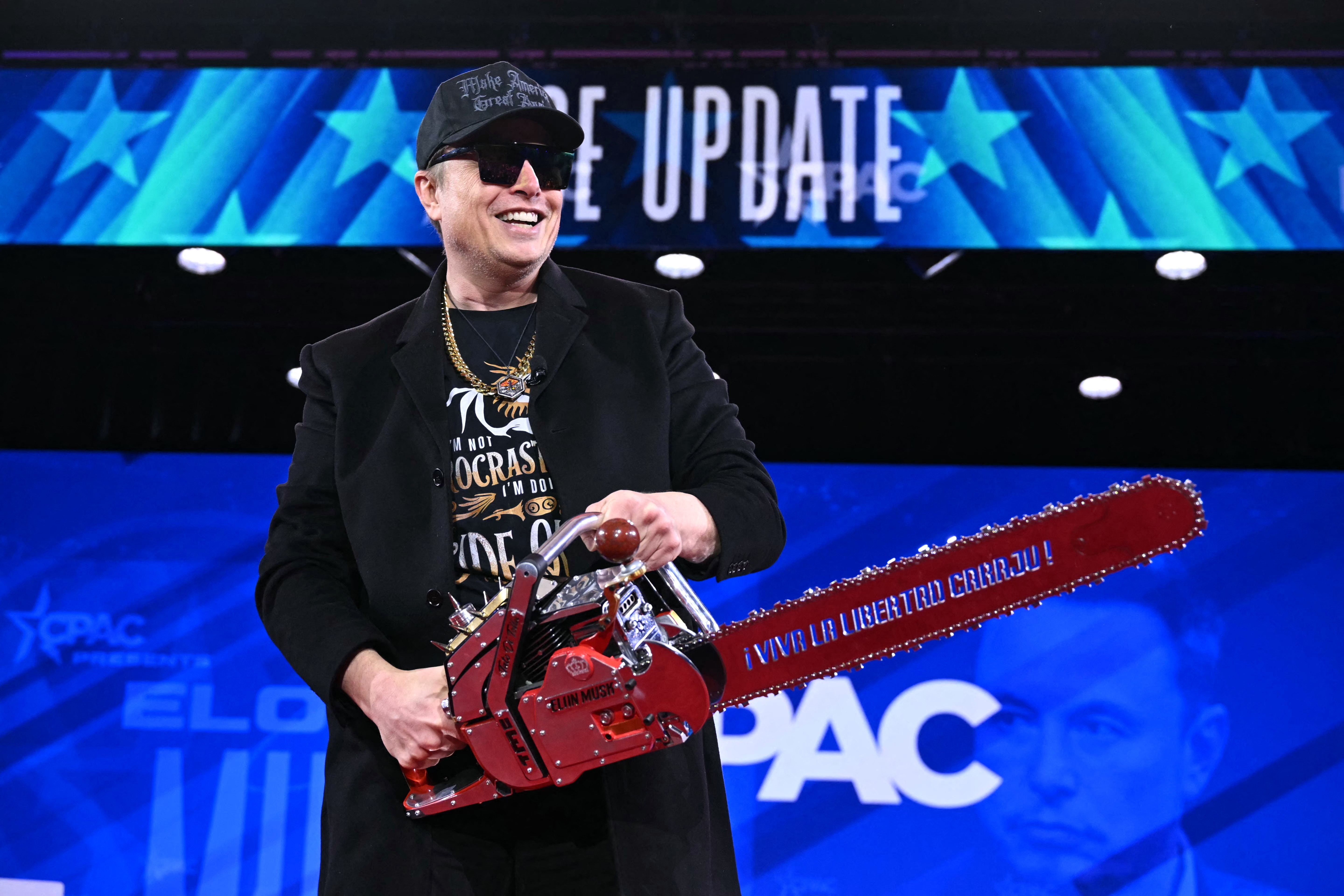 Elon Musk in a suit and hat holding a red chainsaw on stage at an event with "CPAC" signage in the background
