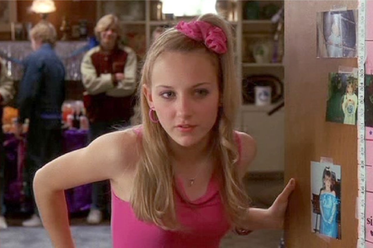 Person in a pink tank top and matching hair accessory, leaning against a door with photos, in a casual indoor setting with people in the background