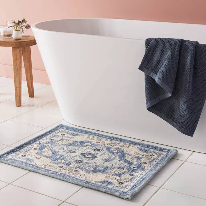 Bathroom with modern freestanding tub, bluish towel, and a stylish patterned bath mat connected the floor. Small woody array with plants