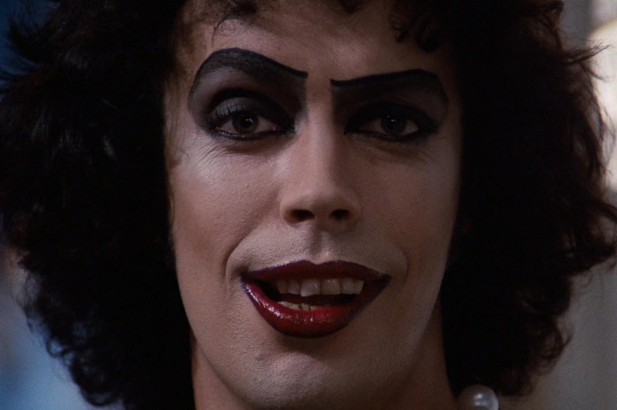 Person with dramatic dark eye makeup and bold red lipstick, wearing pearl necklace. Facial expression is confident and engaging