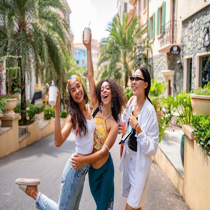 Three people joyfully pose on a sunny street, holding drinks. One wears a headband and tank top, another a floral top, and the third a white jacket