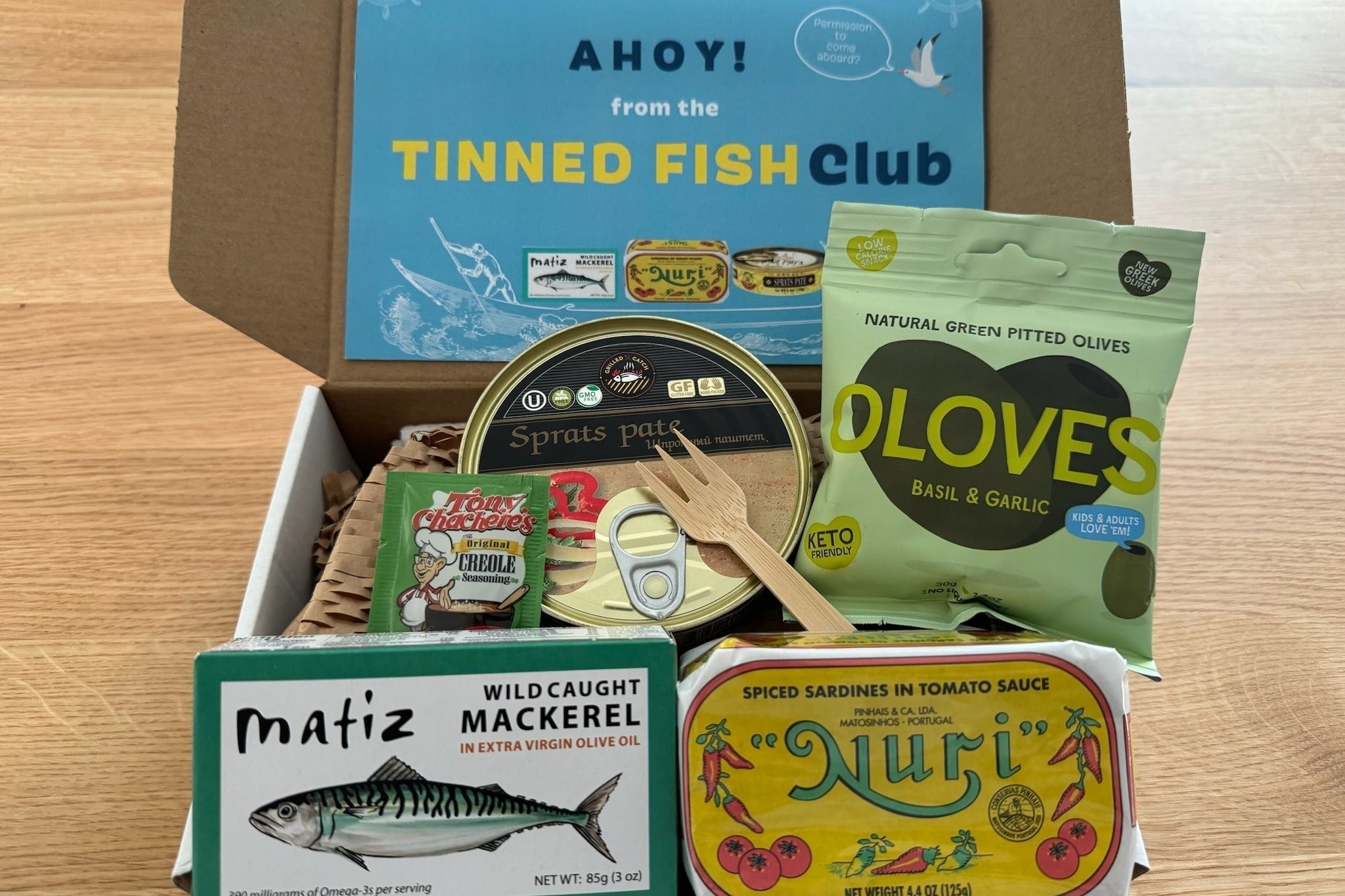 Box of assorted tinned fish and olives with packaging labels visible, part of a subscription promoted by Tinned Fish Club