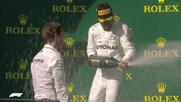 Race car driver celebrates on podium with champagne spray, facing another driver