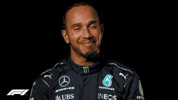 A person in a racing suit with sponsorship logos smiles softly. The suit has a high collar and zipper detailing