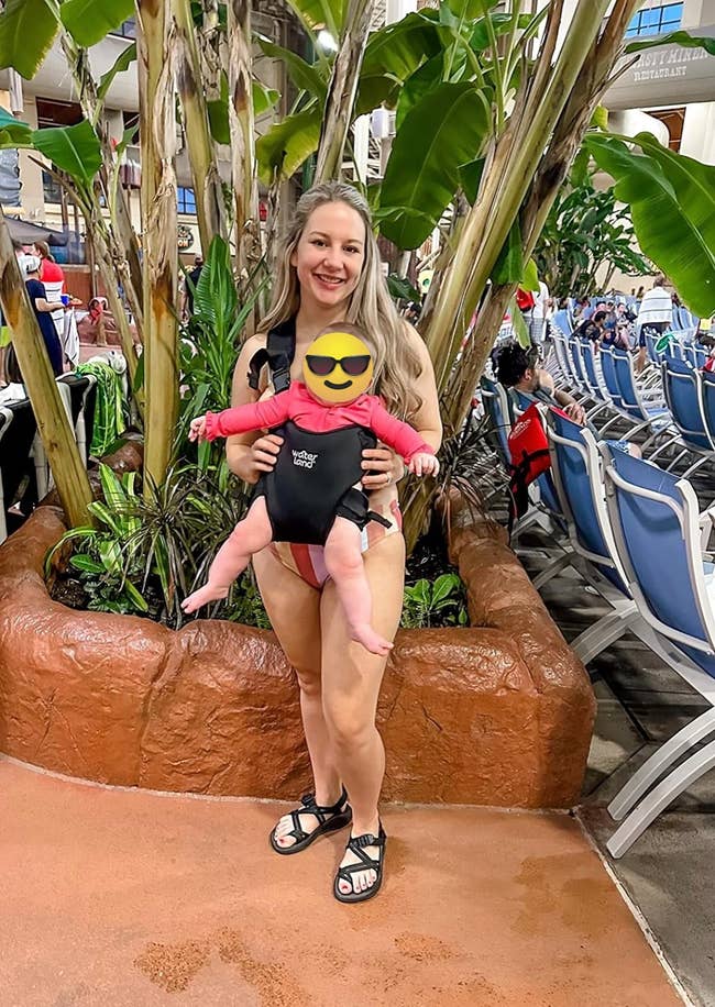 A person smiles while holding a baby in a carrier at an indoor water park; they stand in front of tropical plants and lounge chairs